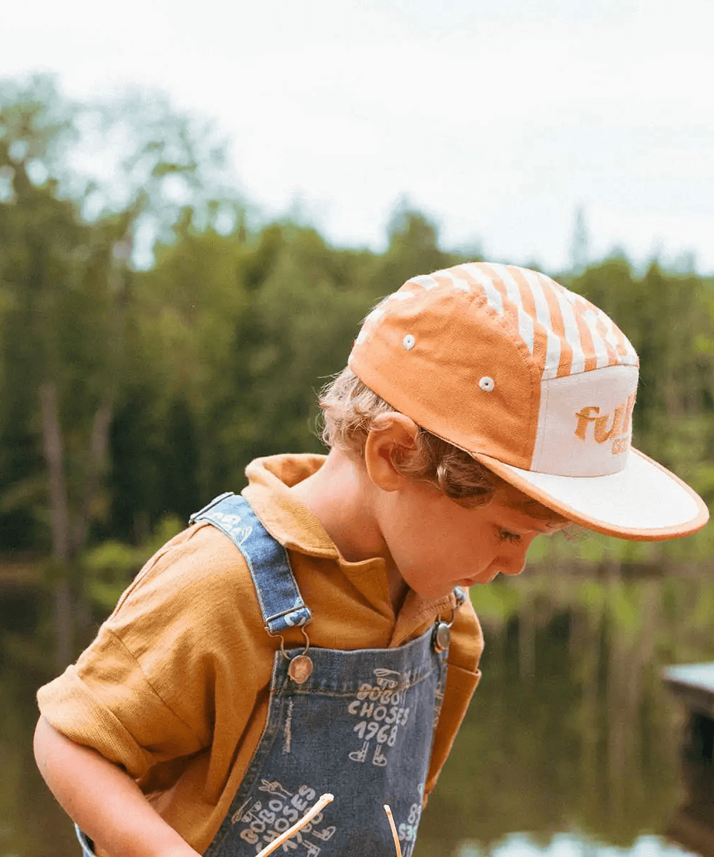 Enfant portant la casquette rayée sable dorée Ki ET LA, vue de profil, en salopette jean au bord d'un lac