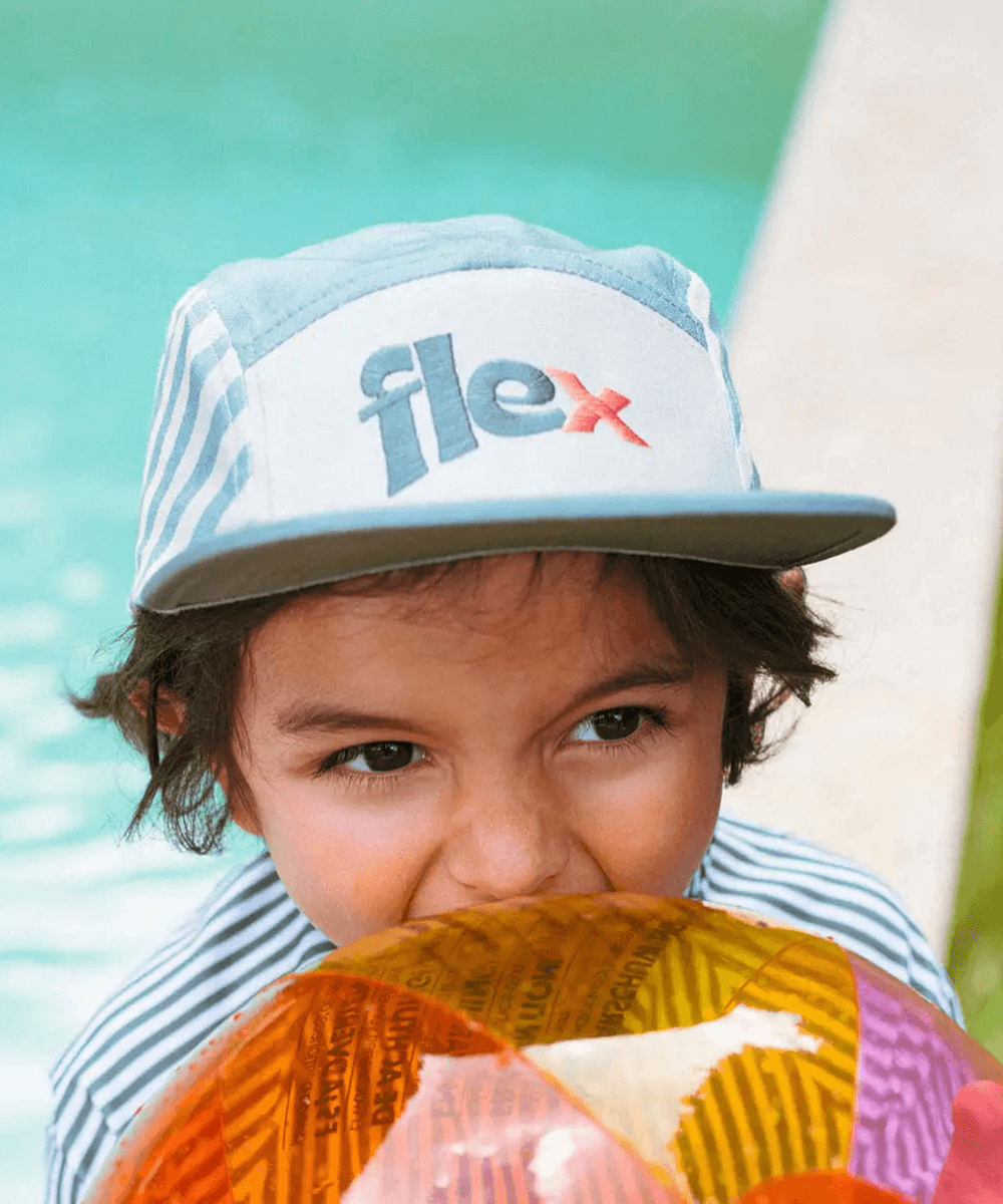 Enfant portant la casquette rayée vert sauge Ki ET LA, vue de face, au bord d'une piscine avec un ballon gonflable devant la bouxhe
