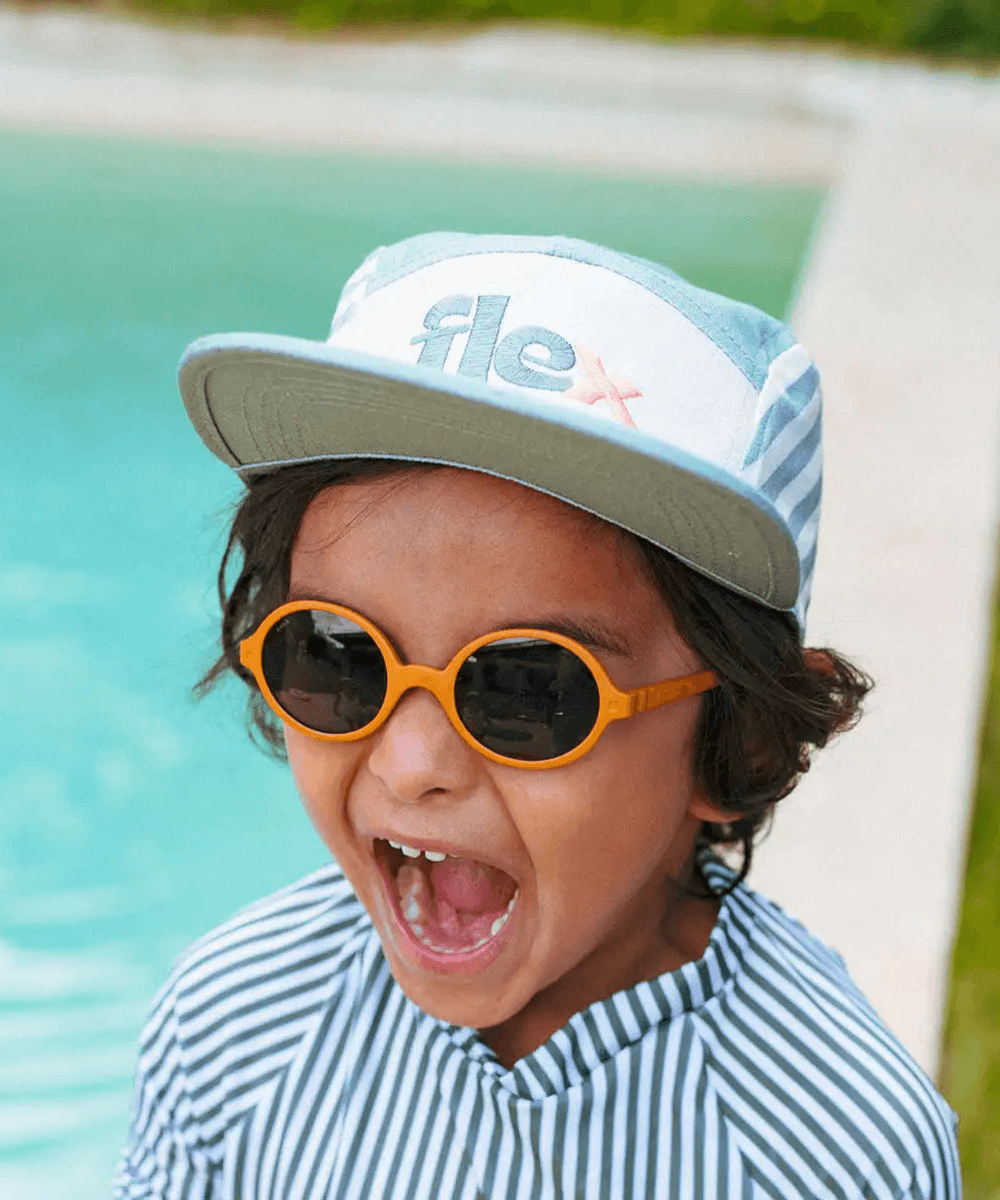 Enfant portant la casquette rayée vert sauge Ki ET LA, vue de face avec des lunettes de soleil, au bord d'une piscine