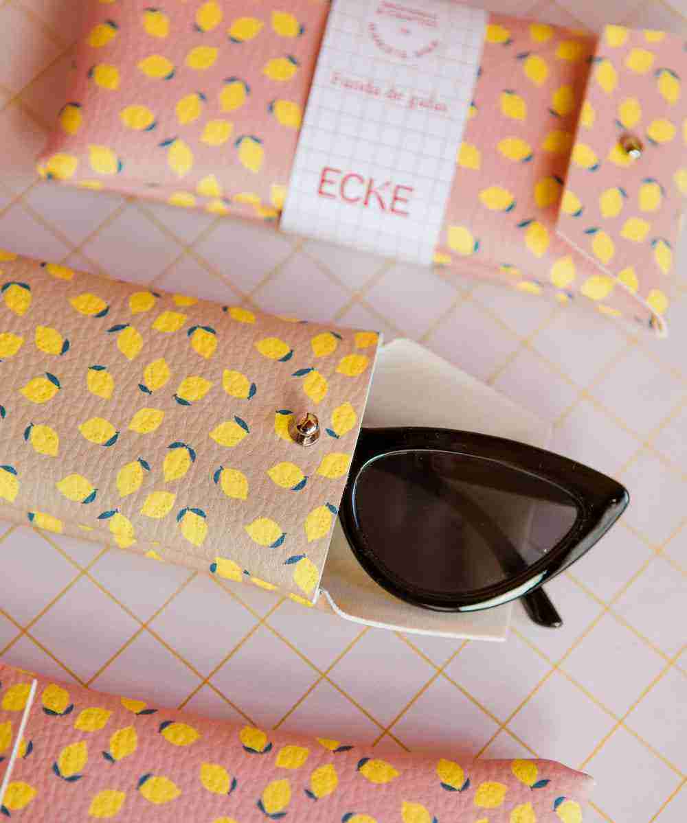 Étui à lunettes ECKE rose à motif citrons, ouvert avec des lunettes noires sur fond quadrillé rose, entouré d’étuis assortis
