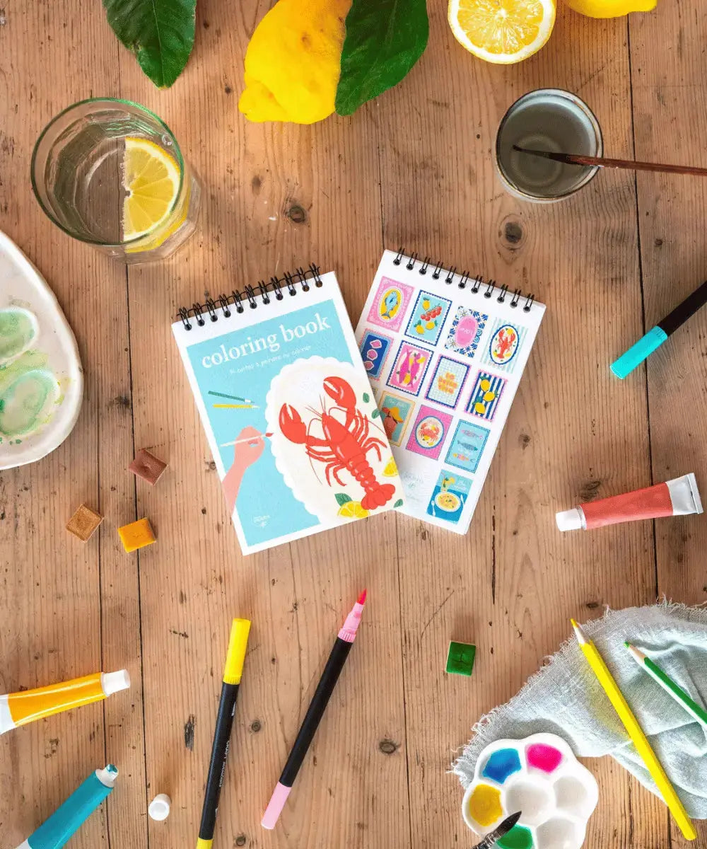 Carnet de coloriage posée sur une table accompagné de tubes de peinture, de feutres et de crayons