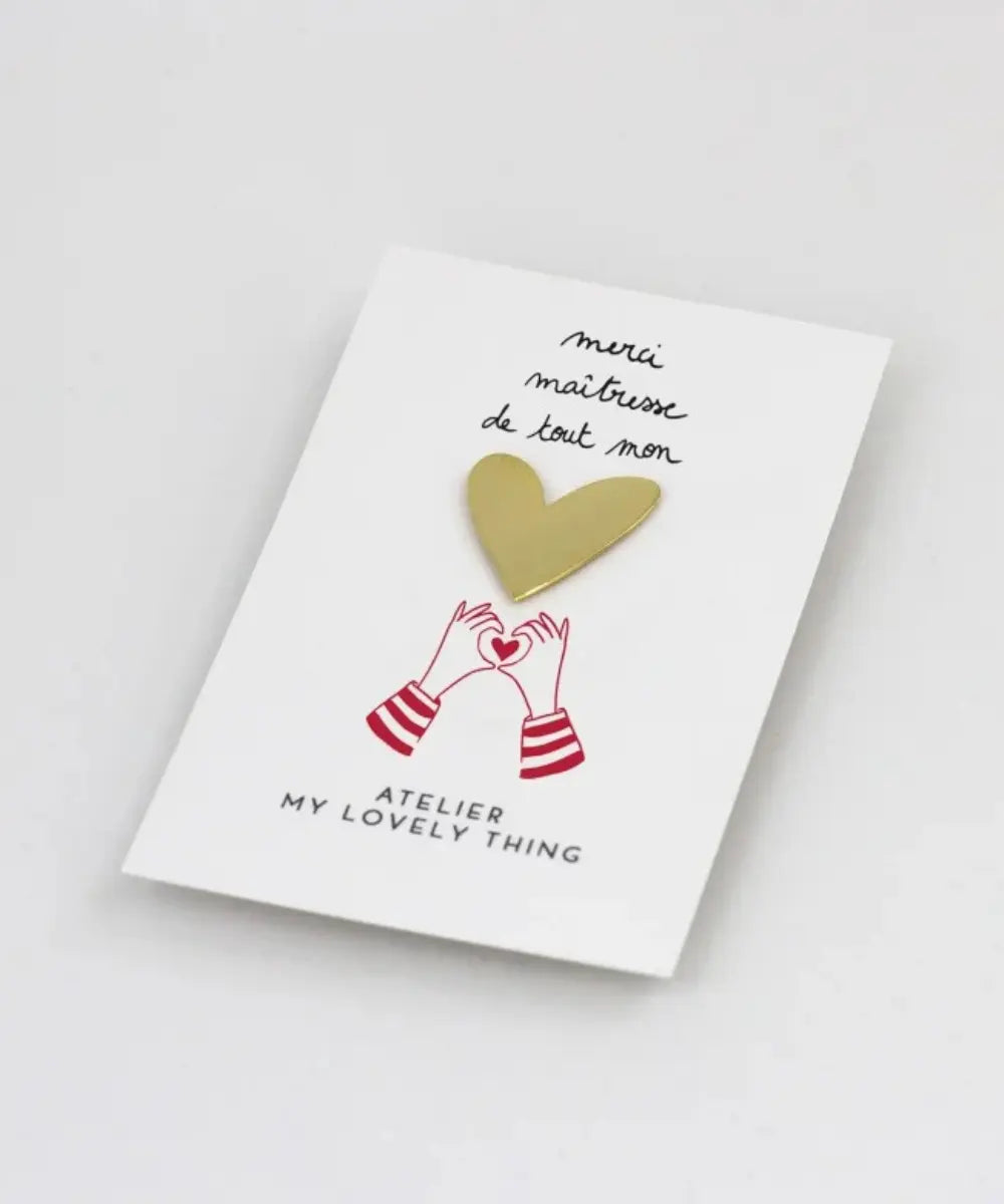 Petit pin's en laiton en forme de cœur doré brillant posé sur sa carte cadeau illustrée Merci Maîtresse.