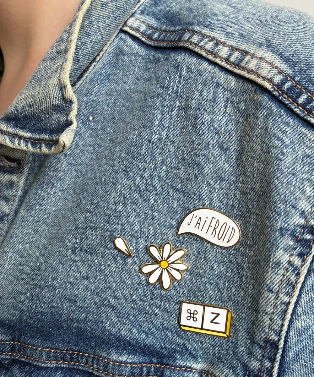 Petit pin's émaillé en forme de marguerite blanche aux contours dorés sur sa carte cadeau.